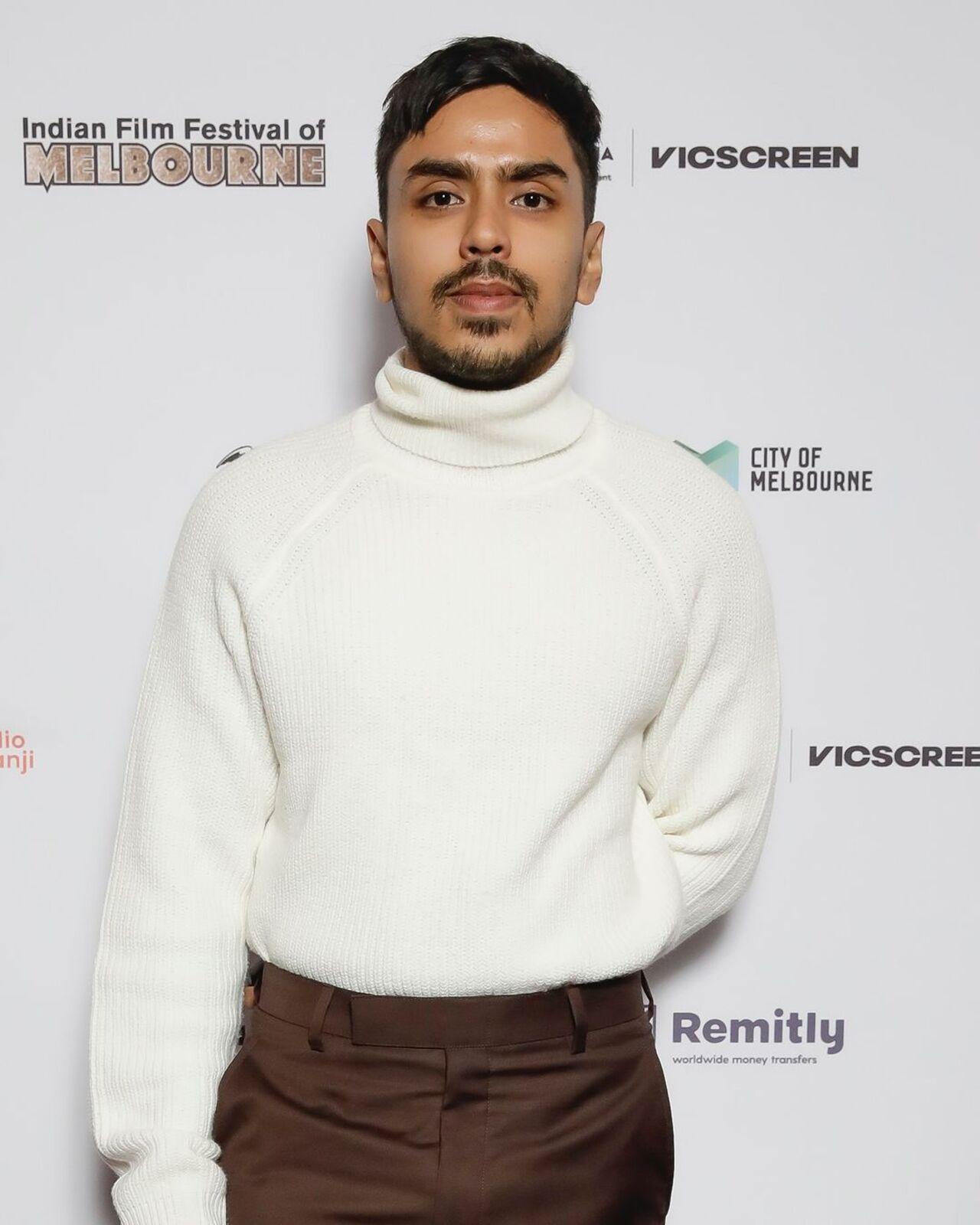 Adarsh Gourav poses on the premiere night red carpet in a white turtleneck sweatshirt and brown pants.