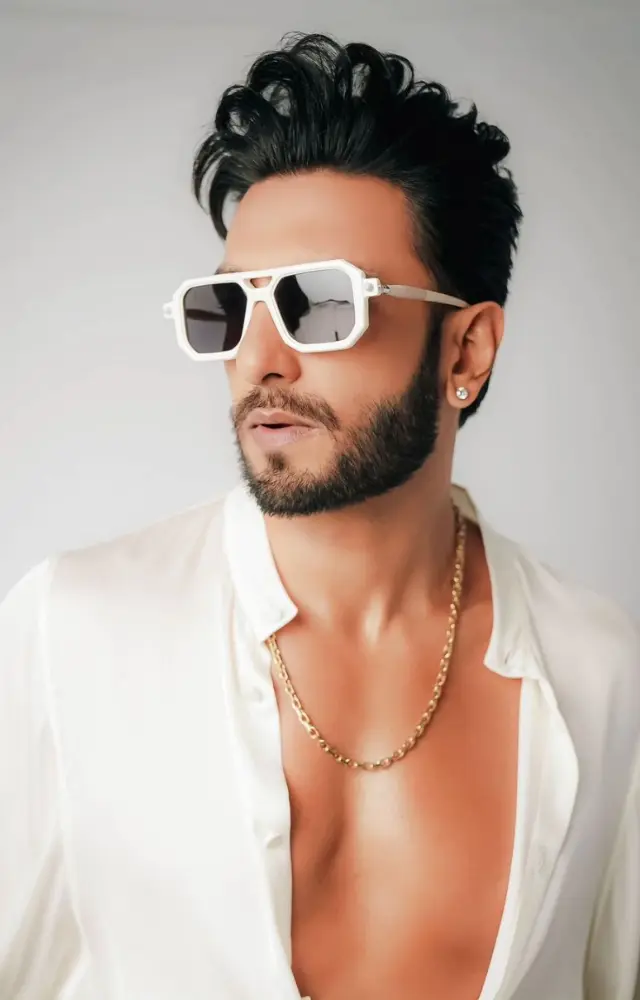 The look is accessorized with white rectangular sunglasses, a thin gold chain necklace, a large, statement watch, and several rings, adding a touch of luxury