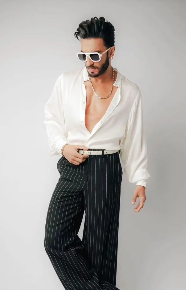 Ranveer features a stylish combination of a deep V-neck white silk shirt paired with high-waisted black pinstriped trousers